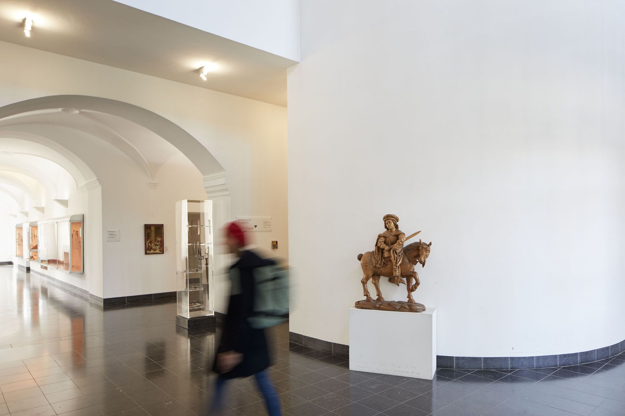 Ein Flur mit einer Statue und Vitrinen. Die Statue aus Holz zeigt einen Reiter mit Schwert und Pferd. In den Vitrinen sind Dolche und alte Schwerter zu erkennen. Über den Flur läuft ein/eine Besucher:in.